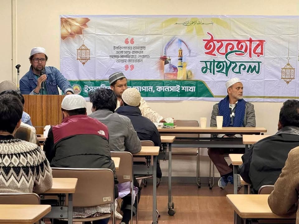 A man wearing a white cap speaks into a microphone at a podium in front of a Bengali-language Iftar Mahfil banner, while a group of men sit at tables listening, including one man wrapped in a scarf seated near the front.