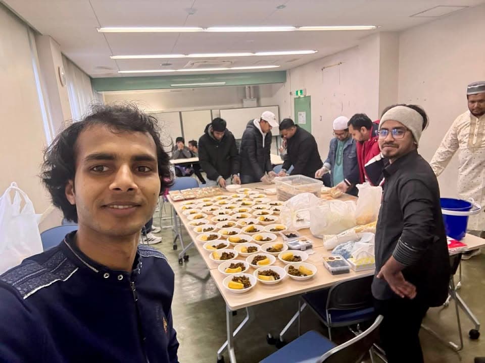 A man takes a selfie in a community hall while others behind him arrange bowls of dates and fruit on long tables for iftar preparation.