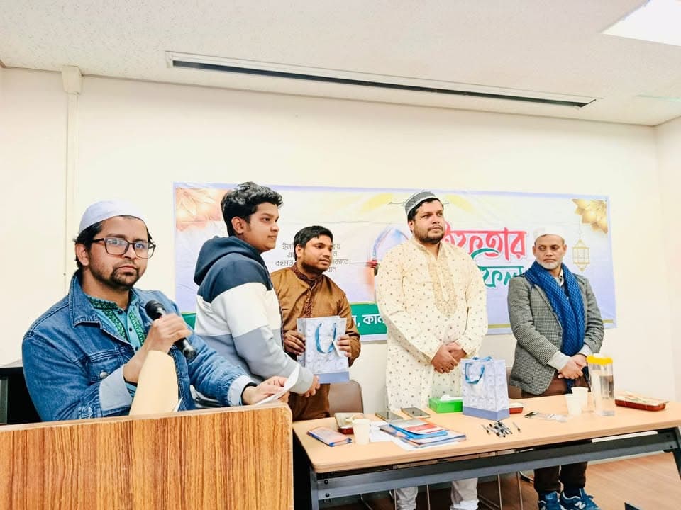 A man in a denim jacket and cap speaks into a microphone at a podium, while four other men stand behind a table holding gift bags and prizes, in front of a banner with Bengali text.