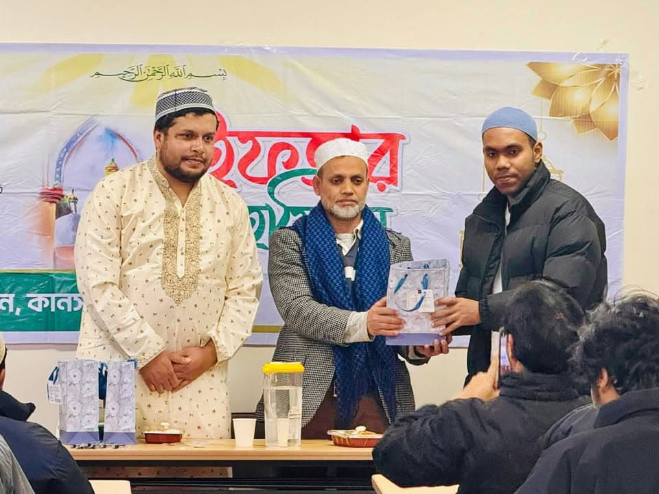 Three men stand in front of a decorated banner with Bengali and Arabic text; one man in a blazer and scarf hands a gift bag to another man in a black jacket, while a man in a white kurta stands beside them, and seated attendees watch in the foreground.