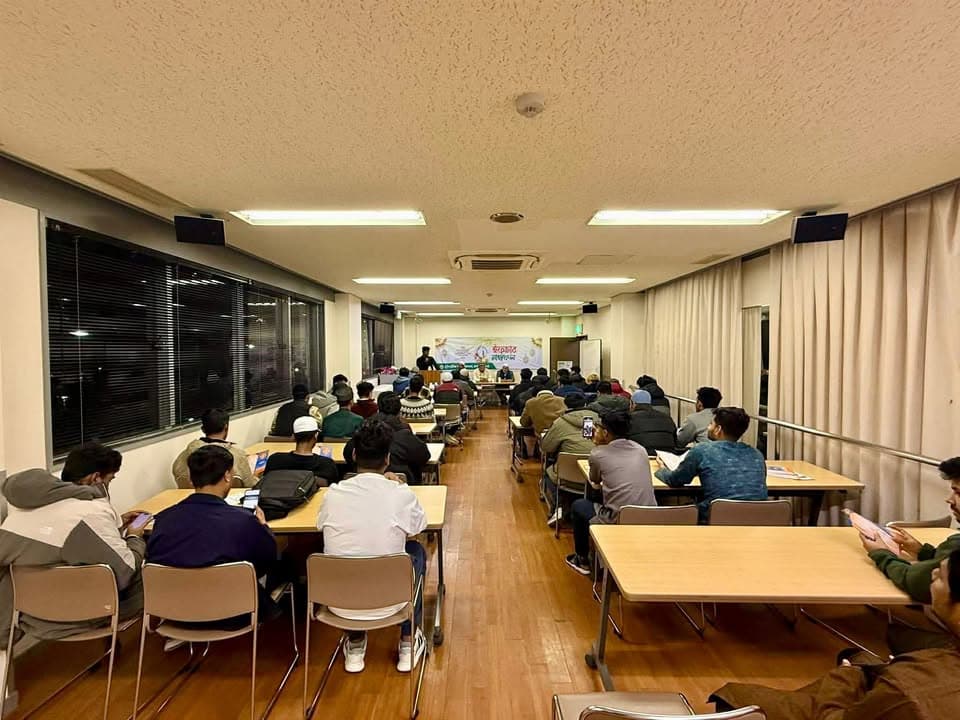 Rows of attendees sit at long tables in a community center hall, listening to speakers seated at a front table decorated with a colorful banner.