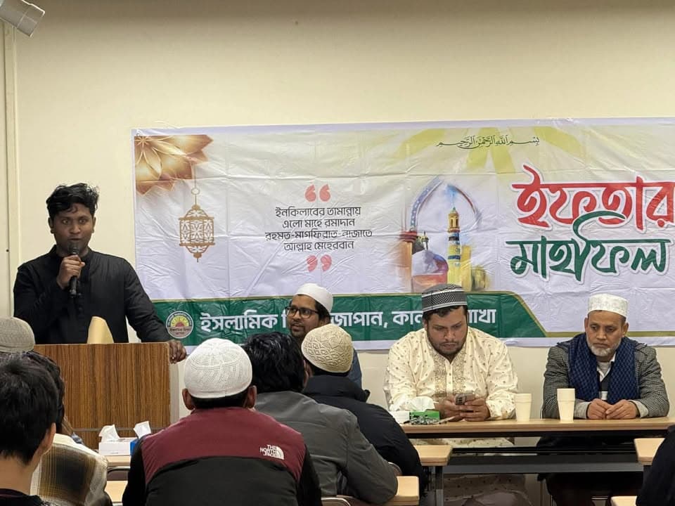 A man speaks into a microphone at a podium beside a large Bengali-language banner reading "Iftar Mahfil," while several men wearing kufi caps sit at tables in front of him listening.
