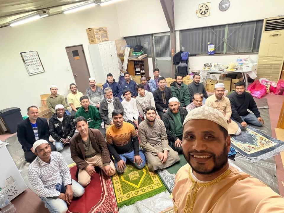 A large group of men, many wearing prayer caps, kneel together on prayer rugs in an indoor community hall, posing for a group selfie taken by a man in a beige robe in the foreground.