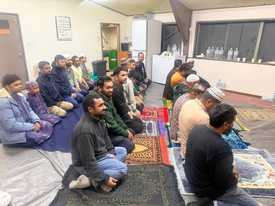 A group of men and boys sit cross-legged on prayer mats and carpets spread across a room's floor, waiting together before iftar, with bottled water and refreshments lined up on the windowsill behind them.