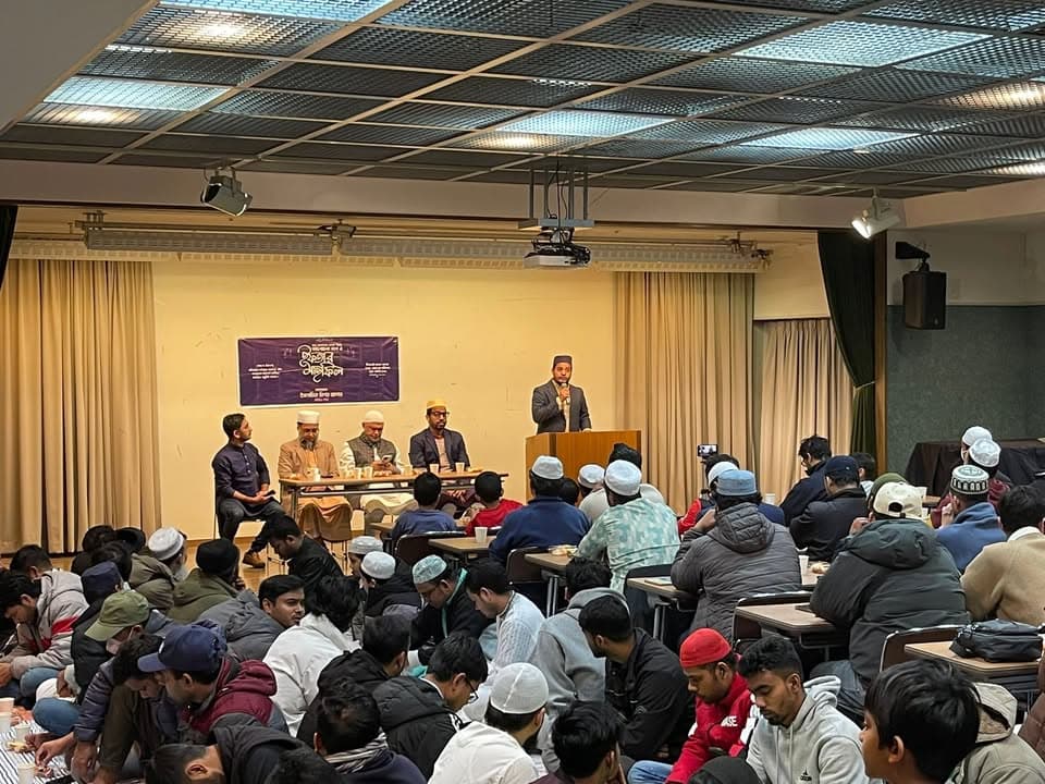 A large gathering of men, many wearing prayer caps, seated on the floor and at tables in a hall, listening to a speaker at a podium in front of a stage with a banner and four seated panelists.