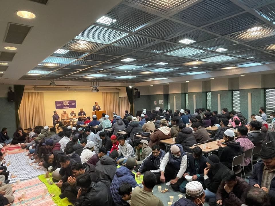 A large hall filled with rows of men sitting on the floor and at tables, waiting to break their fast, while a speaker addresses the gathering from a podium at the front near a banner.