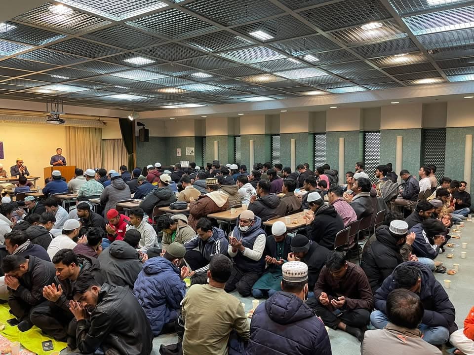 A large hall filled with men seated on the floor and at tables, hands raised in supplication before iftar, while a speaker addresses the gathering from a podium at the front.