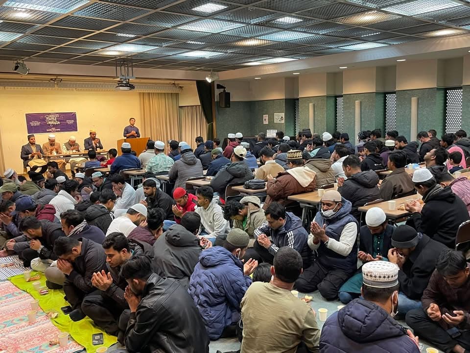 A large hall filled with rows of men seated on the floor and at tables, hands raised in prayer, while speakers sit on a stage in front of a banner in the background.