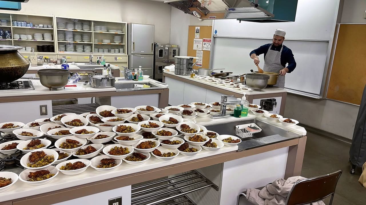 A commercial kitchen counter filled with dozens of small white bowls of cooked food, while a man in an apron and cap stirs a large pot at a stove in the background.