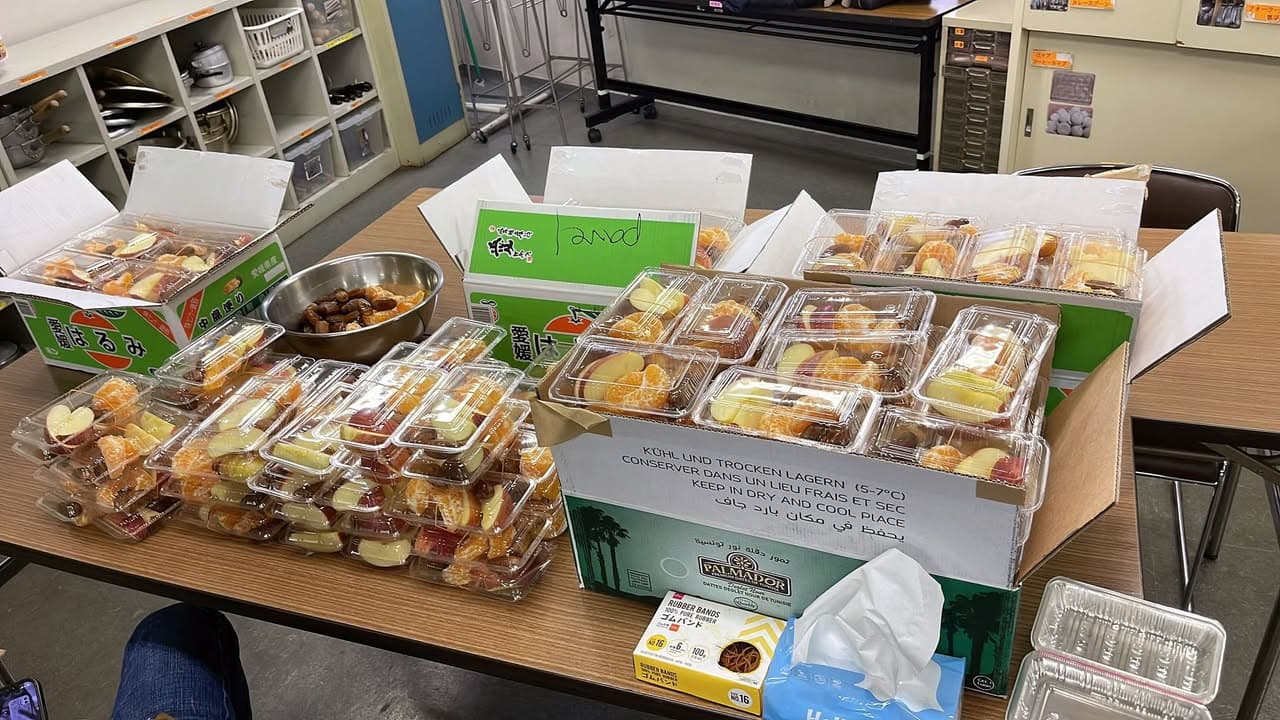 A table filled with small plastic containers of sliced fruit such as mandarin oranges, apples, and dates, along with a metal bowl of dates and open cardboard fruit boxes, prepared indoors for distribution.