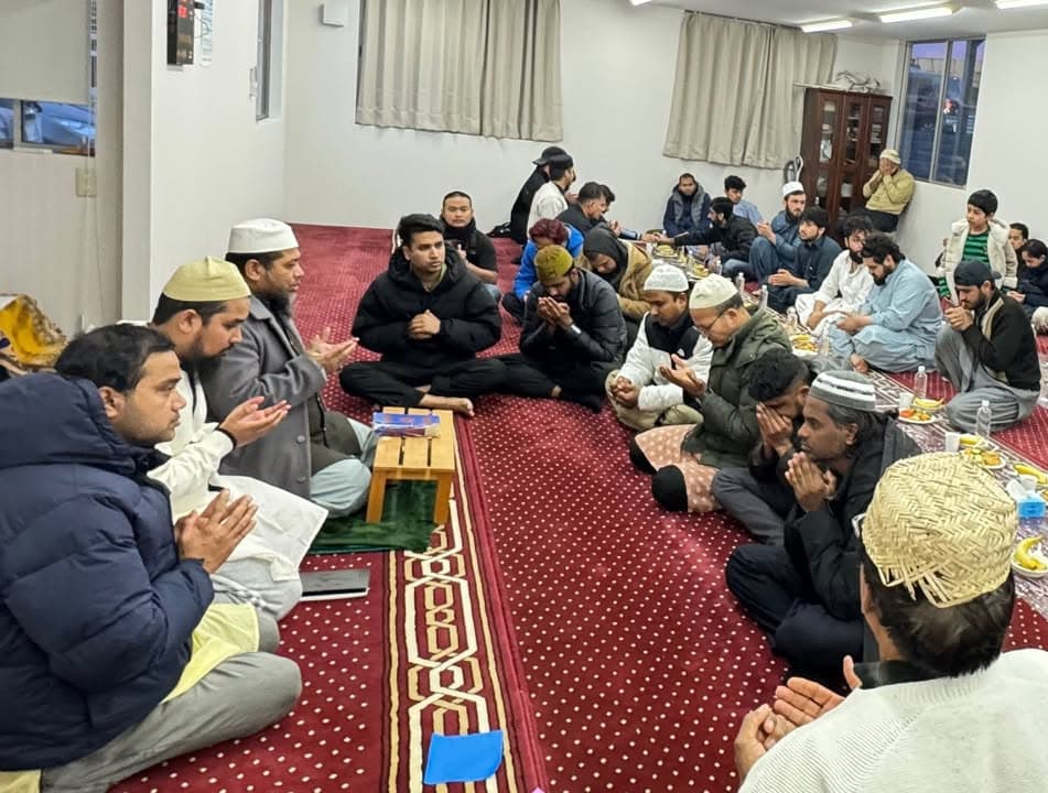 A group of men and boys sit cross-legged on a red patterned carpet inside a mosque hall, hands raised in dua before breaking their fast, with plates of dates and food set out in front of them.
