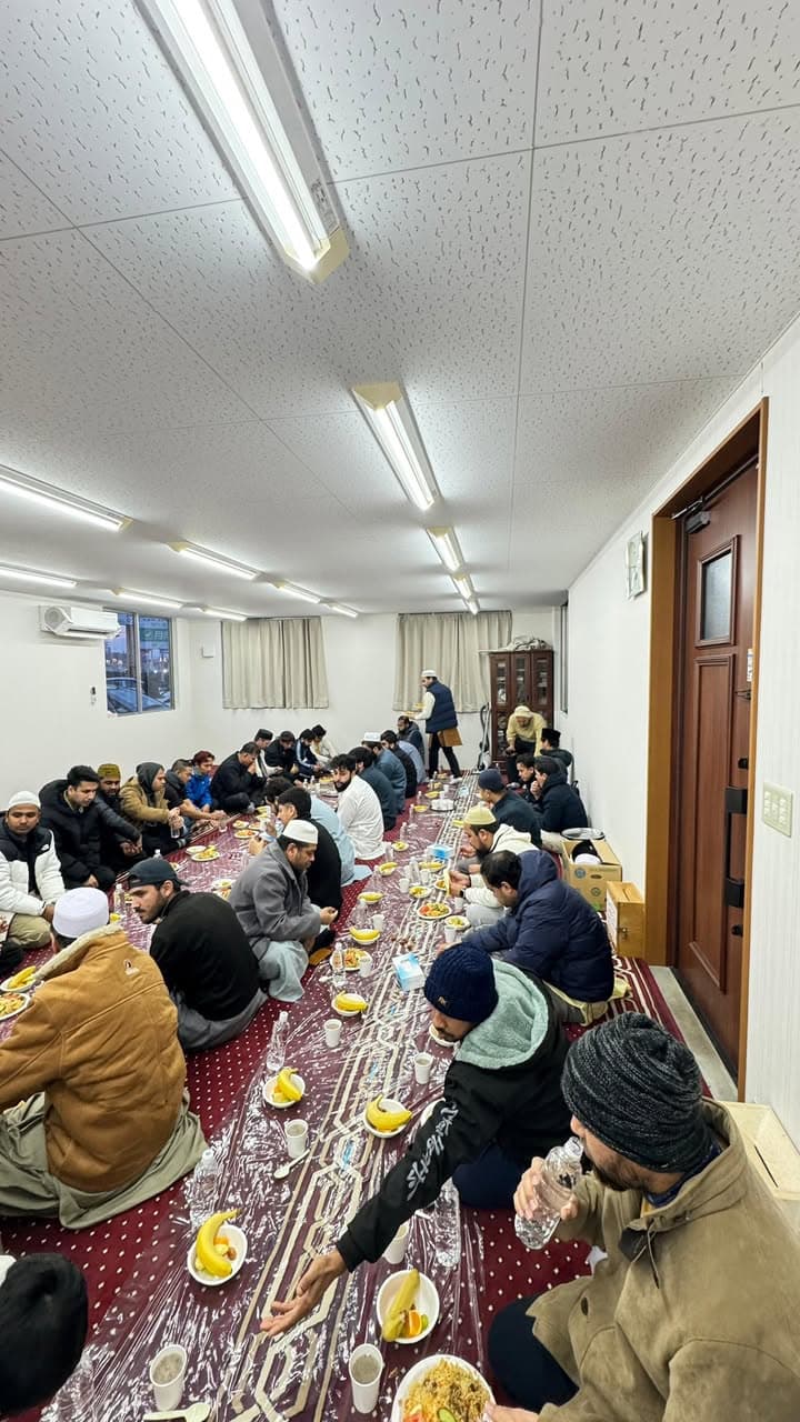 Men sit in two long rows on a plastic-covered carpet inside a mosque hall, sharing plates of food, fruit, and bottled water for an iftar meal.