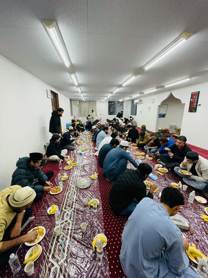 Rows of men sit cross-legged on plastic sheeting on a red carpeted floor inside a mosque hall, eating plates of food with bananas, cups and water bottles laid out along a long communal dining spread under bright ceiling lights.