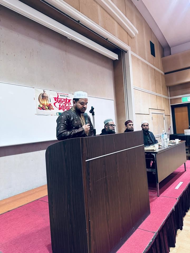 A man in a cap and embroidered kurta speaks into a microphone at a podium on a stage, with three other men in caps seated at a table behind him and a Bengali-language Iftar Mahfil poster on a whiteboard in the background.