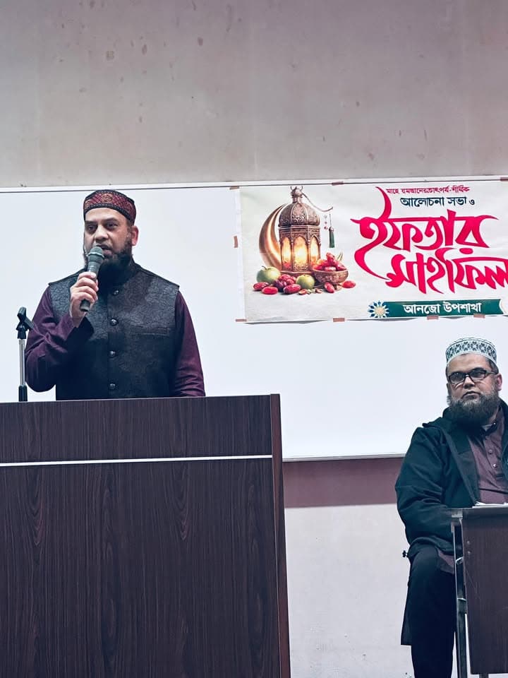A man wearing a maroon vest and embroidered cap speaks into a microphone at a podium, with another man seated nearby beside a colorful banner written in Bengali announcing an Iftar Mahfil.