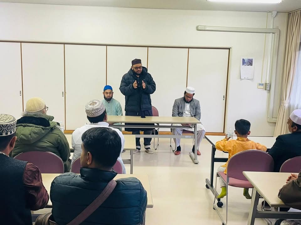 A man wearing a cap and winter jacket stands and speaks while gesturing with his hands, in a Japanese-style tatami community room with seated attendees including men and a child at desks arranged in rows.