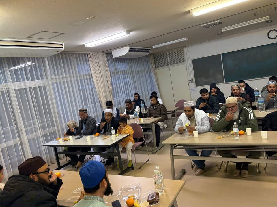 Men, women and children sit at classroom-style tables eating oranges, dates and snacks to break their fast, with sheer curtained windows and a blackboard in the background.
