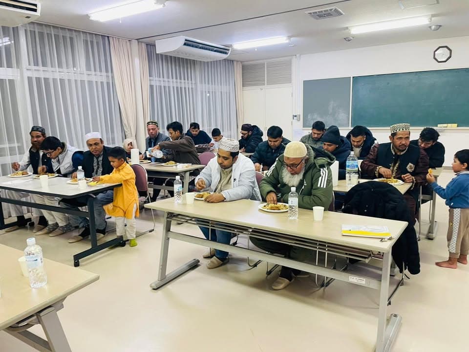 Men, women, and children sit at rows of tables in a classroom-like hall, eating plates of food together, with bottled water and cups on the tables.