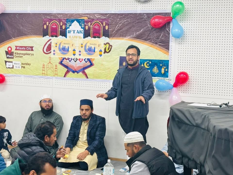 A man stands speaking to a seated audience in a community hall decorated with balloons and a banner reading "Iftar Mahfil," with participants sitting on the floor around him during a gathering.