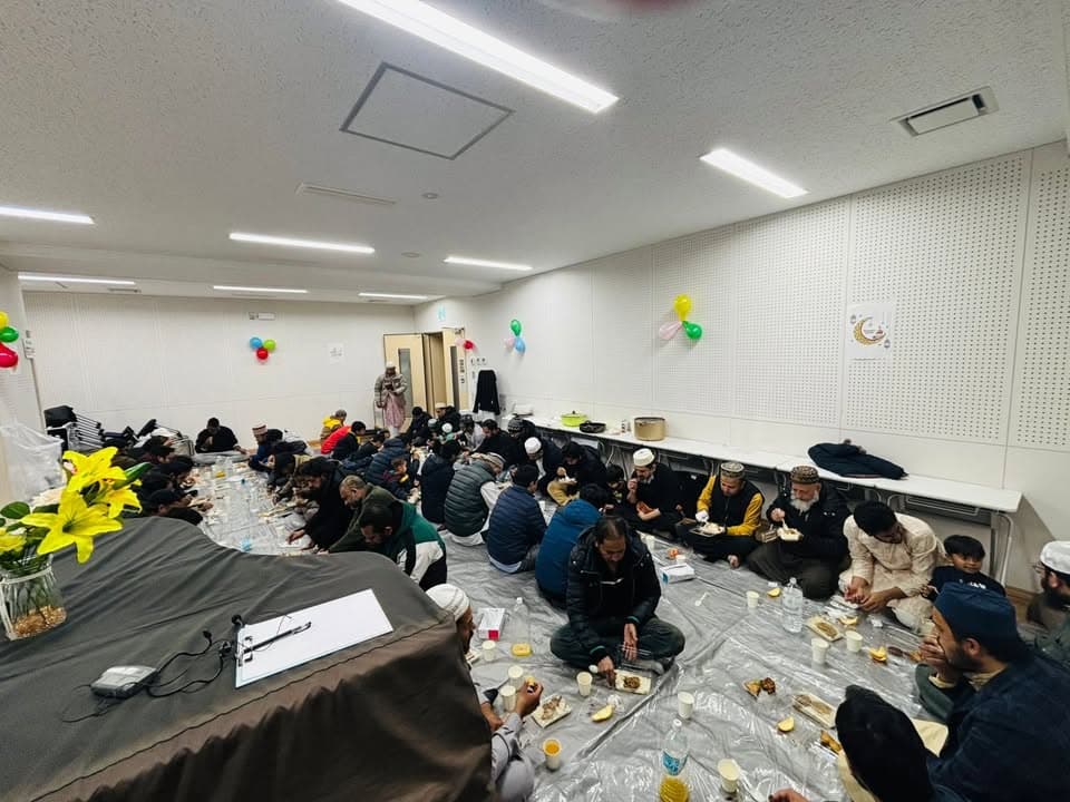A large group of men and boys sit together on plastic sheeting spread across a hall floor, eating a meal from plates and cups, with balloons and a crescent moon decoration on the walls behind them.