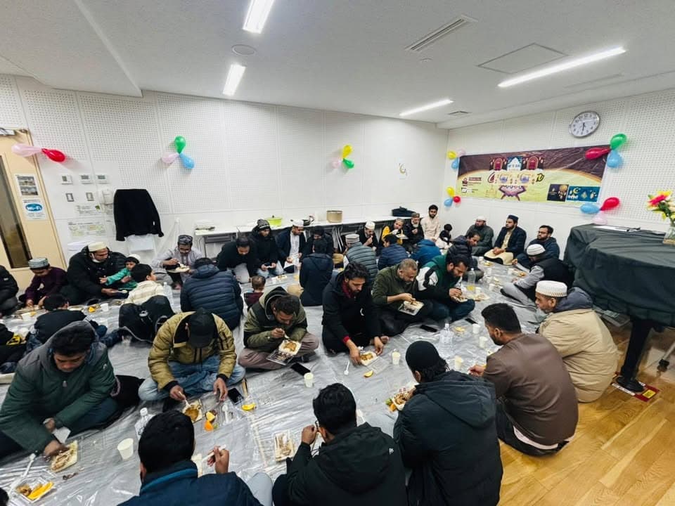 A large group of men sit on plastic sheeting spread across the floor of a hall decorated with colorful balloons and a Ramadan banner, eating a meal together from plates and containers.