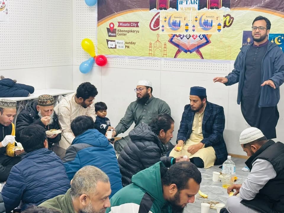 A group of men and a child sit together on the floor of a community hall, eating and talking, while a man in traditional dress stands and speaks at right, with a colorful Iftar Mahfil banner and balloons decorating the wall behind them.