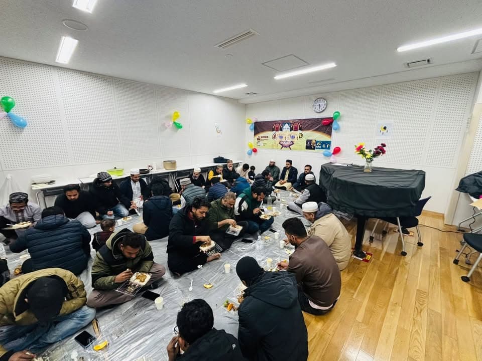 A large group of men sit together on plastic sheeting spread over a wooden floor in a hall decorated with colorful balloons and a Ramadan banner, eating a meal from trays and plates.