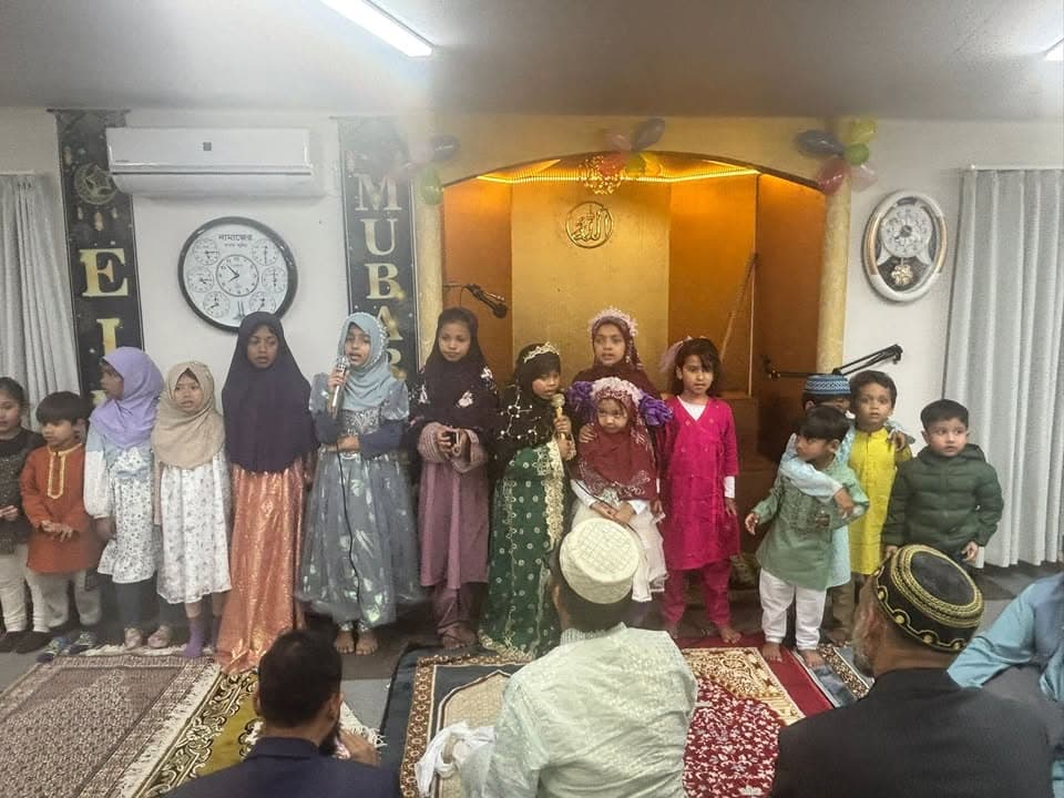 A group of children in colorful traditional outfits stand together holding a microphone and speaking or singing in front of a decorated mosque mihrab, while adults sit on prayer rugs facing them.