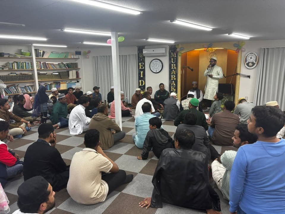 A large group of men and boys sit cross-legged on a checkered carpet inside a mosque hall decorated with "Eid Mubarak" lettering and balloons, listening to a man speaking into a microphone at the front.