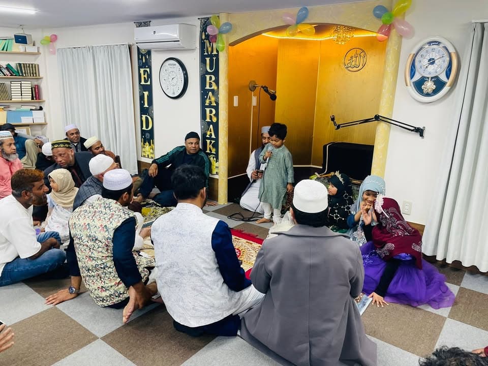 Inside a mosque decorated with balloons and an "EID MUBARAK" banner, a young child holds a microphone and speaks in front of the mihrab while a seated audience of men, women and children listens.