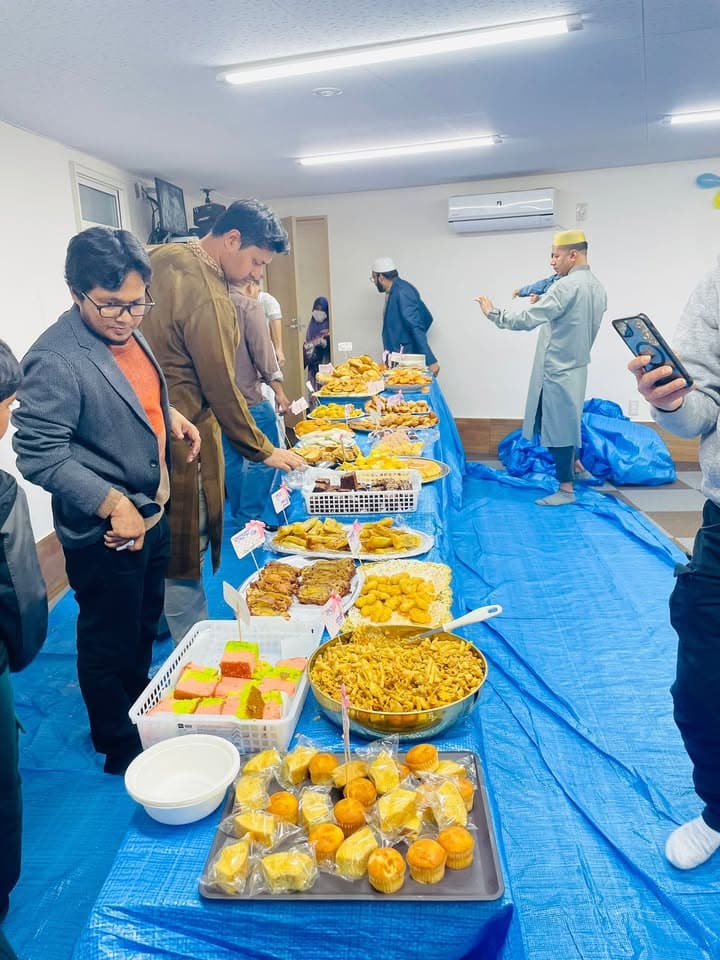 People stand along a long table covered with a blue tarp, laden with a variety of traditional pitha sweets, fried snacks, cupcakes and dishes laid out on plates and trays inside a mosque hall.