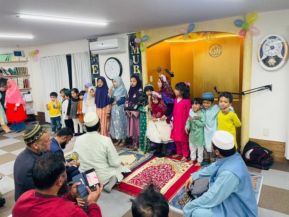 Children and women in colorful traditional dress stand lined up in front of a mosque's mihrab decorated with balloons and "Eid Mubarak" letters, with seated men and photographers in the foreground of the prayer hall.