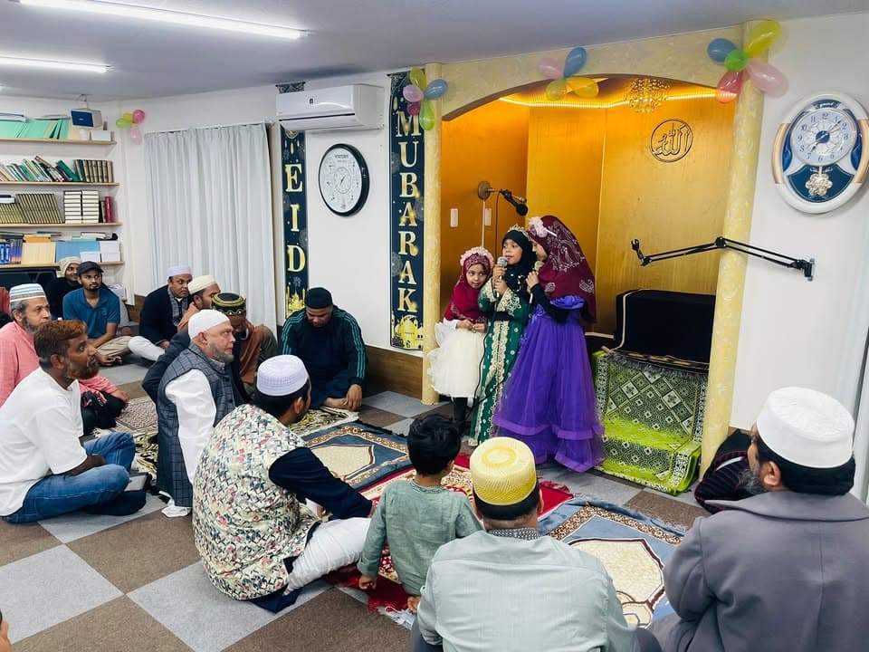 Three young girls in colorful traditional dresses stand near a decorated mihrab holding a microphone, performing in front of a seated audience of men and boys in a mosque hall decorated with "Eid Mubarak" banners and balloons.