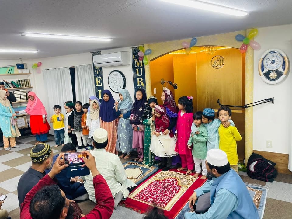 Children and young women in colorful traditional dress stand together in front of the mosque's golden mihrab, decorated with balloons and a "MUBARAK" banner, while men seated on the floor take photos with their phones.