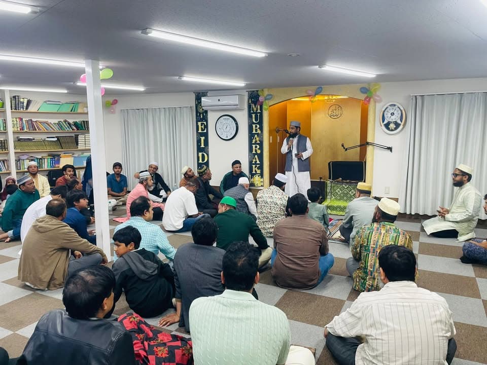 A man speaks into a microphone at the front of a prayer hall decorated with "Eid Mubarak" signage and balloons, while a large group of men and boys sit on the carpeted floor listening.