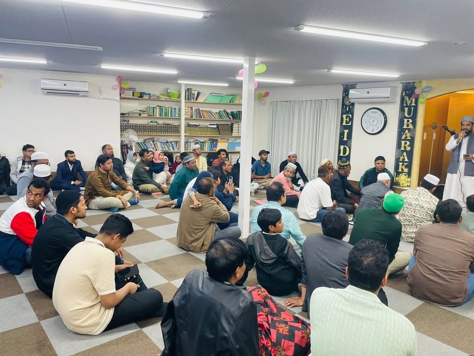 A large group of men sit cross-legged on a checkered floor inside a mosque hall decorated with "EID MUBARAK" banners and balloons, listening to a man speaking into a microphone at the front of the room.