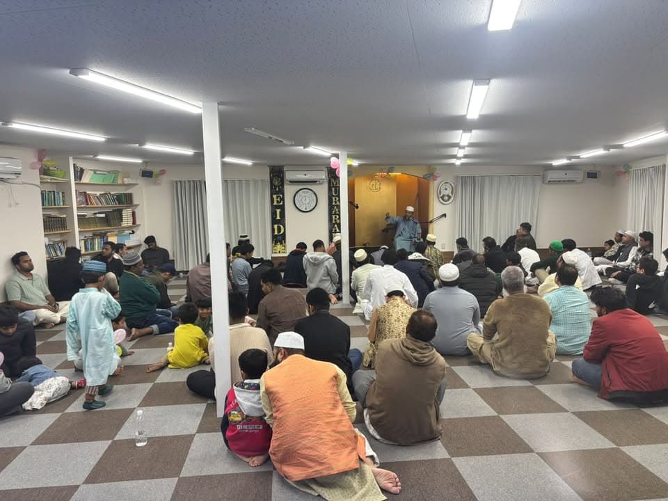 A large group of men and children sit cross-legged on a checkered mat floor inside a mosque hall, listening to a man speaking at a lectern in front, with an "EID MUBARAK" banner and balloons decorating the wall.