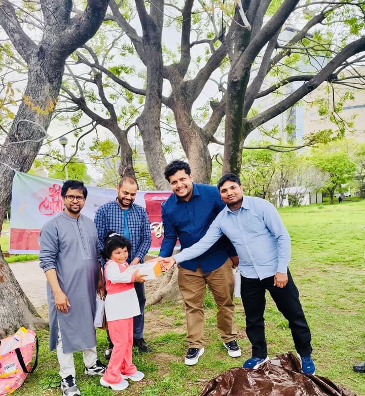 Four men and a young girl stand together under a large tree in a park, with one man handing a small box to the girl, and a red banner with Bengali text visible in the background.