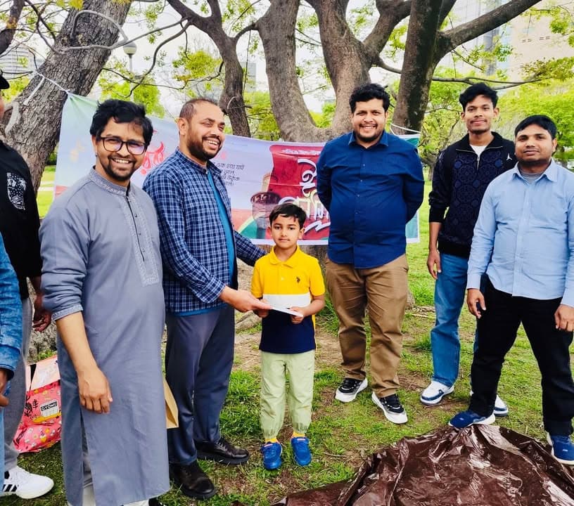 A group of men and a young boy stand outdoors under trees in a park, with a man handing an envelope to the boy in front of a colorful Bengali-language banner.