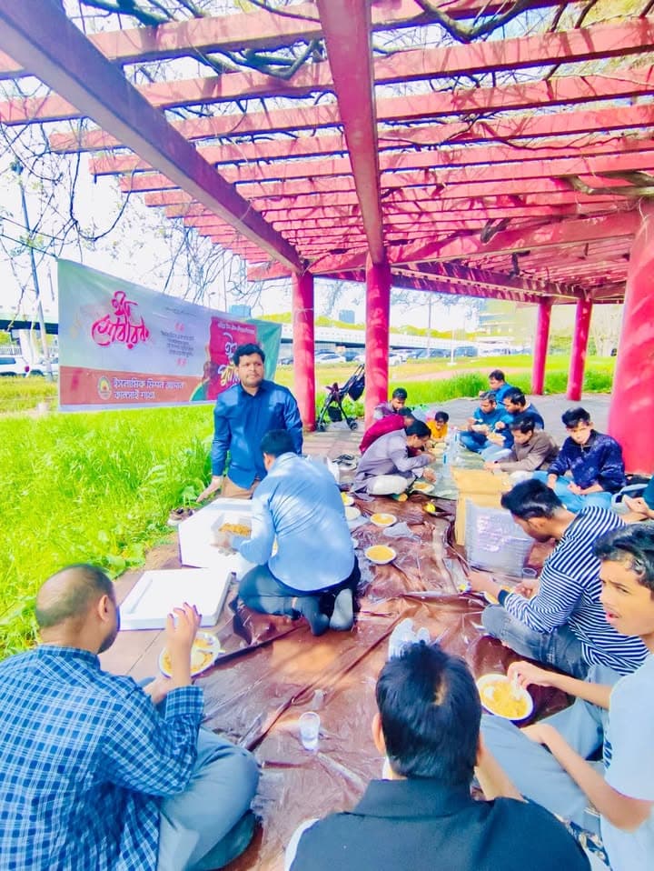 A group of men and boys sit on plastic sheeting under a red pergola in a park, eating from plates while a man in a blue shirt serves food from a box, with a Bengali-language banner set up in the grass nearby.