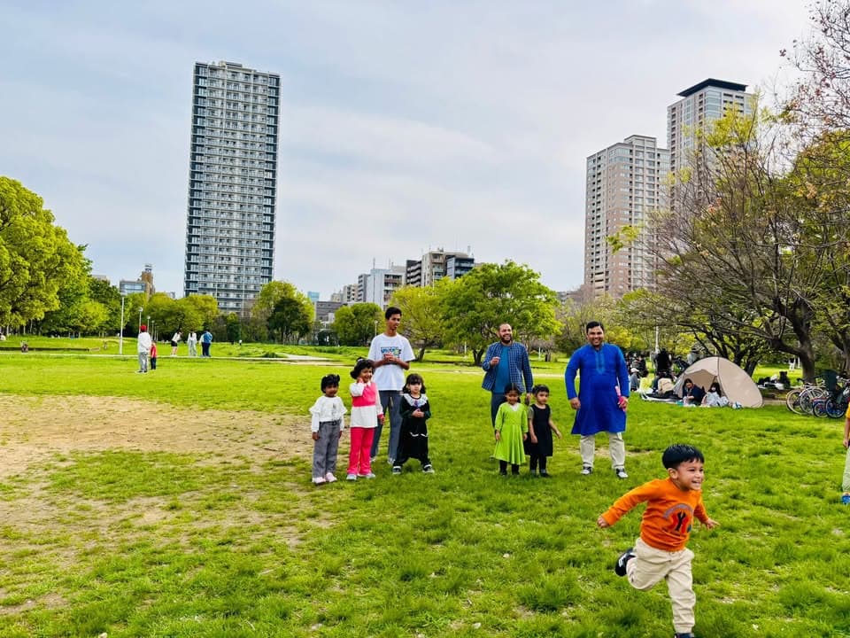 A family group of children and men stand and play on a grassy park lawn with high-rise buildings in the background, as a young boy runs across the grass in the foreground.