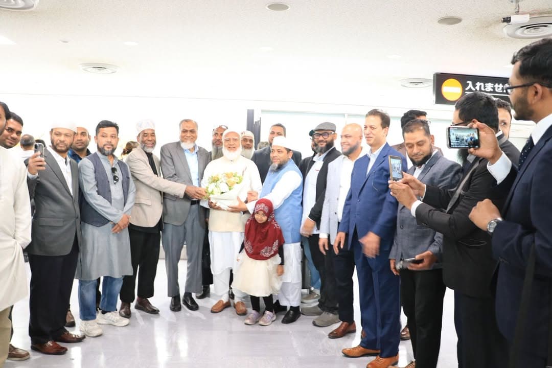 A group of men in formal and traditional attire gather at Narita Airport's arrival hall, presenting a bouquet of flowers to an elderly guest of honor in white robes, with a young girl in a red headscarf standing beside him and others taking photos with phones.