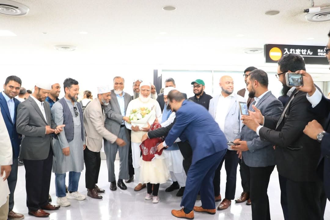 A group of men in an airport arrival hall gather around an elderly man in white robes who is receiving a bouquet of flowers from a young child, while others film and photograph the moment on their phones.
