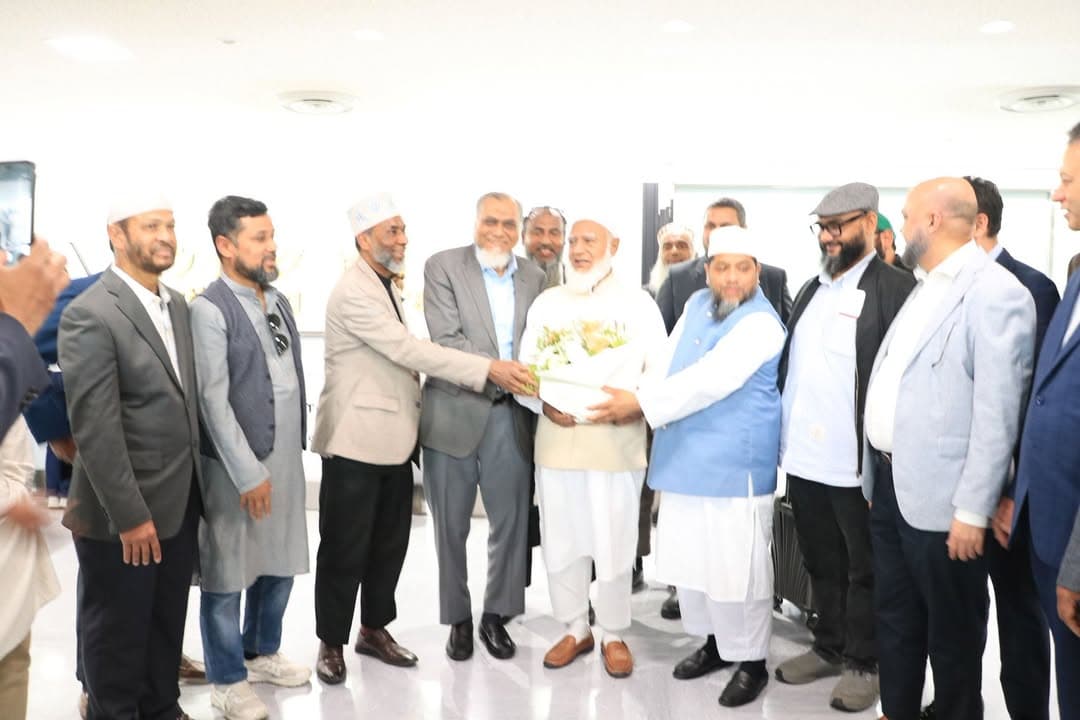 A group of men in suits and traditional attire, including several wearing white caps, gather in an airport terminal as one man presents a bouquet of flowers to an elderly man in white robes and a white cap.