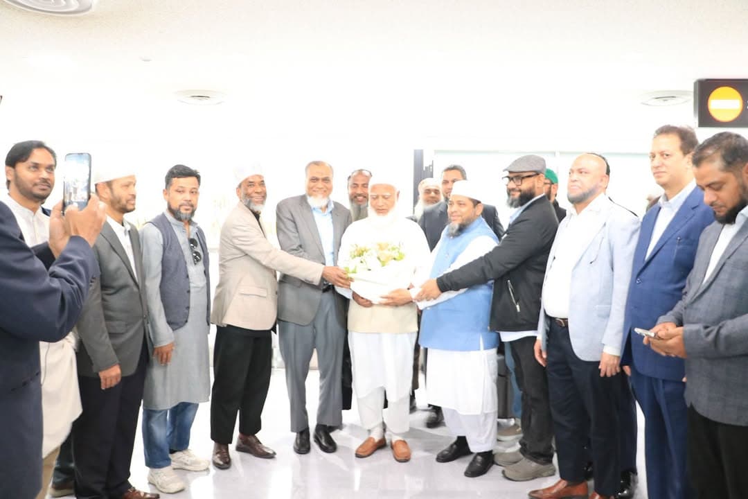 A group of men in formal and traditional attire stand together indoors at an airport, with one man in the center holding a bouquet of white flowers while others shake his hand, and a man on the left taking a photo with his phone.
