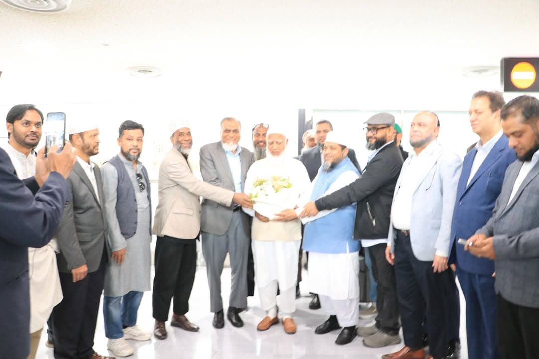 A group of men in an airport terminal presenting a bouquet of flowers to a guest wearing a white cap and traditional dress, while others take photos with a smartphone.