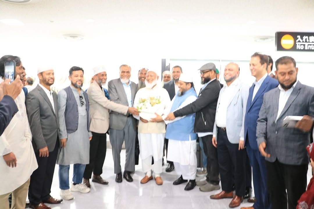 A group of men in suits and traditional dress stand together at an airport arrival area, with one man in white holding a bouquet of flowers as another man reaches out to greet him, while a bystander records the moment on a phone; a "No Entry" sign is visible in the background.