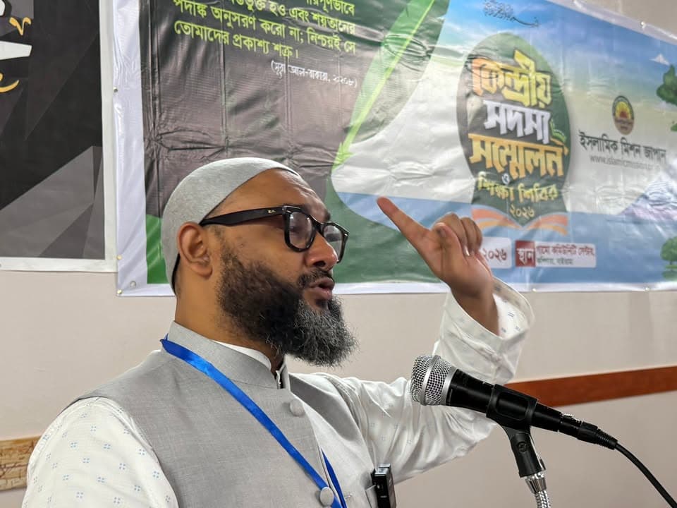 A bearded man wearing glasses and a white kufi cap speaks passionately into a microphone, raising his index finger, with a Bengali-language banner reading "Central Members' Conference and Education Camp 2026" and an Islamic Mission Japan logo behind him.