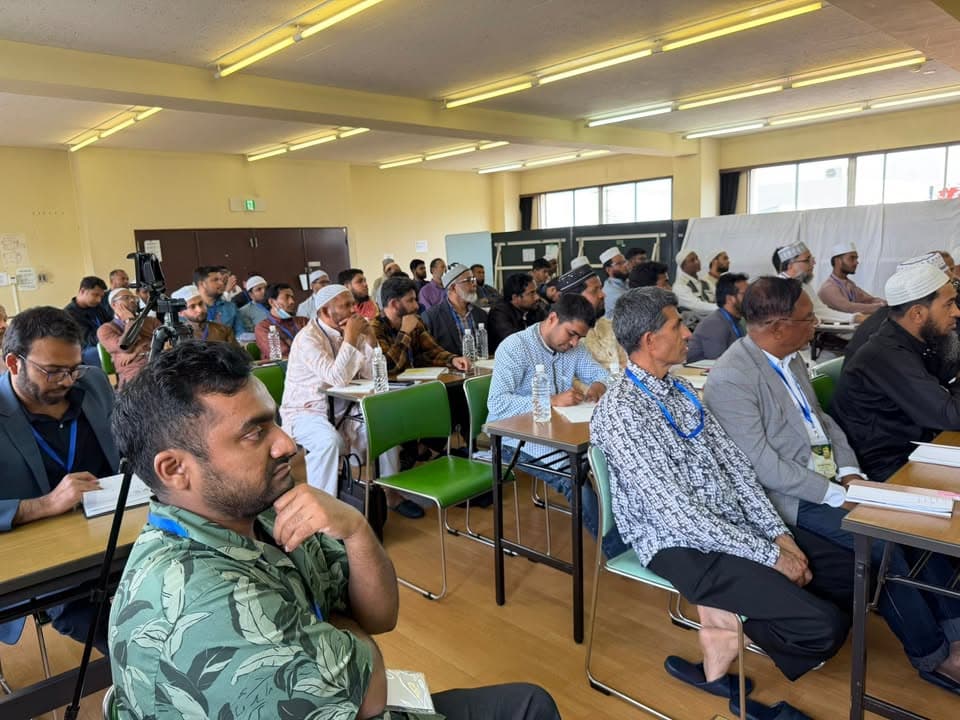 Rows of men, some wearing prayer caps, sit at desks in a conference hall listening attentively to a speaker off-camera, with a camera on a tripod set up in the aisle.