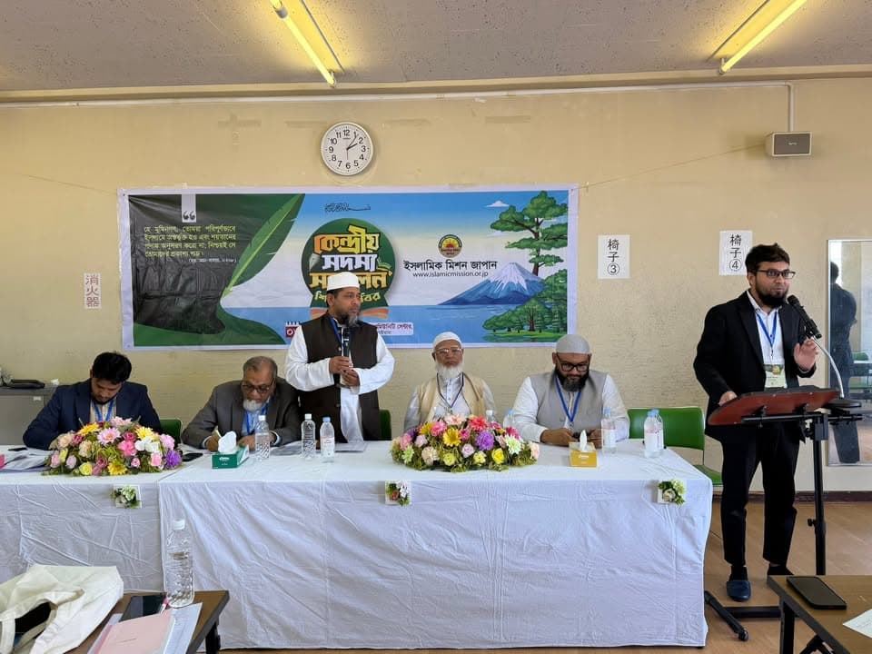 Five men sit and stand behind a long table draped in white cloth with floral arrangements, one man speaking into a microphone while another man stands at a podium to the right, in front of a banner in Bengali and the Islamic Mission Japan logo on a beige wall with a clock.